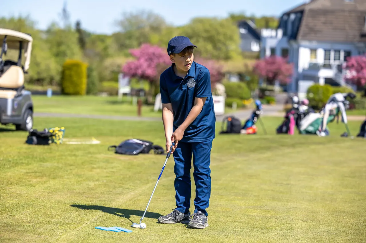 Ein Jugendlicher mit Golfschläger bereitet sich auf einen Putt auf dem Golfplatz vor. Im Hintergrund sind weitere Golfer und ein Golfmobil zu sehen.