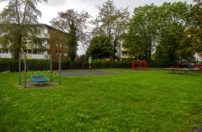 Spielplatz der Regenbogenschule mit einer blauen Netzschaukel, einem roten Klettergerüst und Bäumen vor Wohnhäusern.