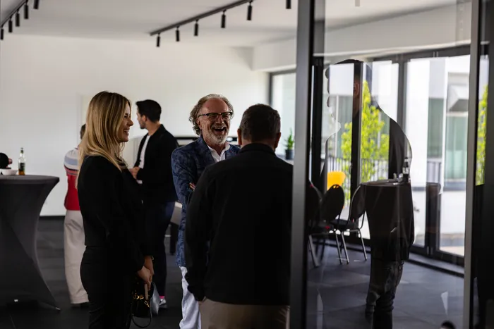 Ein lächelnder Mann mit Brille und blauem Blazer unterhält sich angeregt mit zwei Personen in einem hellen Raum. Im Hintergrund sind weitere Menschen und ein Blick nach draußen zu sehen.