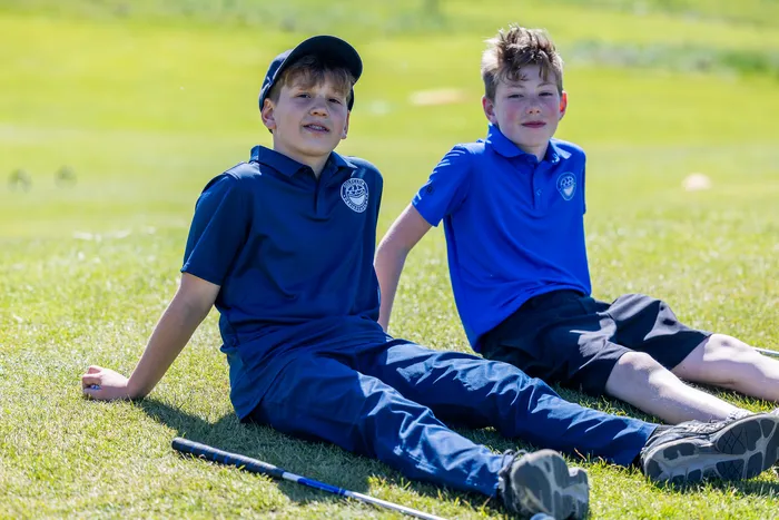 Zwei Jungen sitzen auf einer grünen Wiese, beide tragen blaue Poloshirts mit einem Logo und dunkle Shorts. Im Hintergrund ist eine Golflandschaft zu sehen.