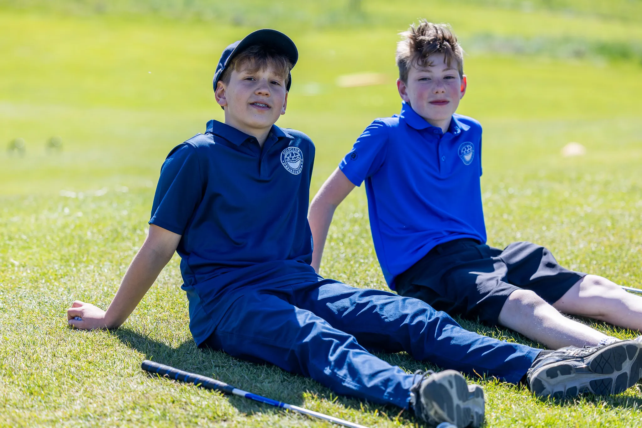 Zwei Jungen sitzen auf einer grünen Wiese, beide tragen blaue Poloshirts mit einem Logo und dunkle Shorts. Im Hintergrund ist eine Golflandschaft zu sehen.