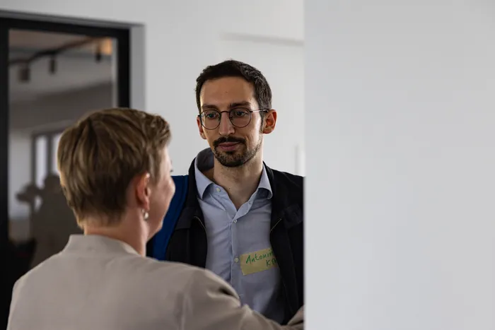 Ein Mann mit Brille und Kurzhaarfrisur blickt zur Seite, während er mit einer Person im Gespräch ist, deren Rücken dem Betrachter zugewandt ist. Er trägt ein Namensschild mit der Aufschrift „Antonim K“.