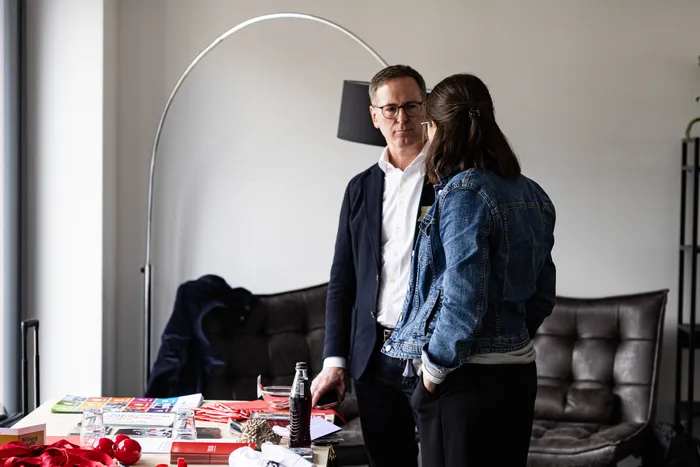 Ein Mann in einem dunklen Anzug und eine Frau mit Jeansjacke stehen nebeneinander vor einem Tisch mit Broschüren und einer Wasserflasche. Im Hintergrund eine große Bogenlampe und ein Sofa.