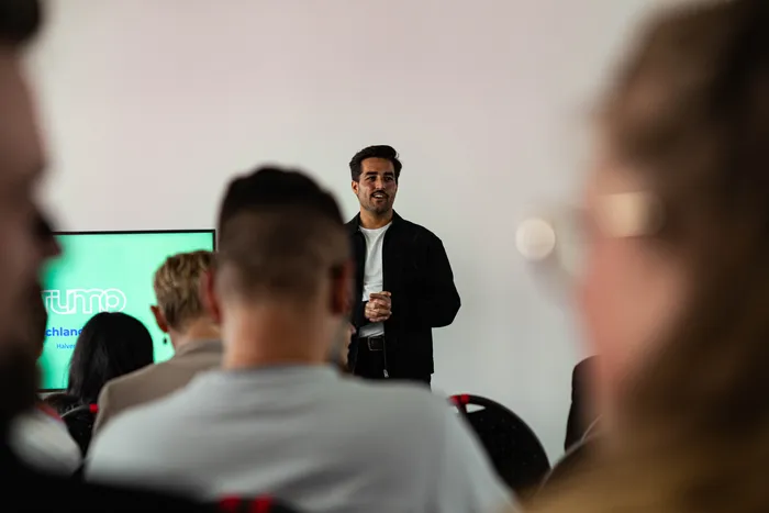 Ein Mann mit dunklen Haaren hält vor einer Gruppe von Menschen eine Präsentation. Im Hintergrund ist eine Präsentation mit dem Schriftzug 