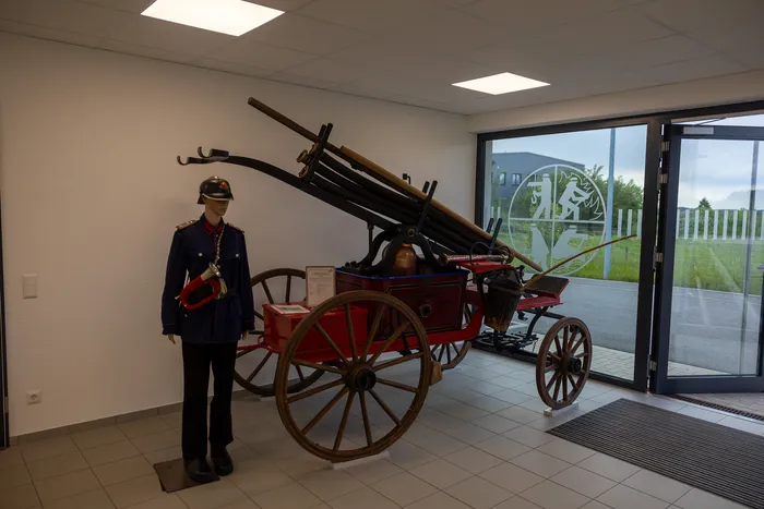 Ein roter, historischer Feuerwehrwagen steht in einem hellen Raum neben einer Schaufensterpuppe in Uniform. Im Hintergrund ist durch eine Glastür ein Gebäude mit einem Logo zu sehen.