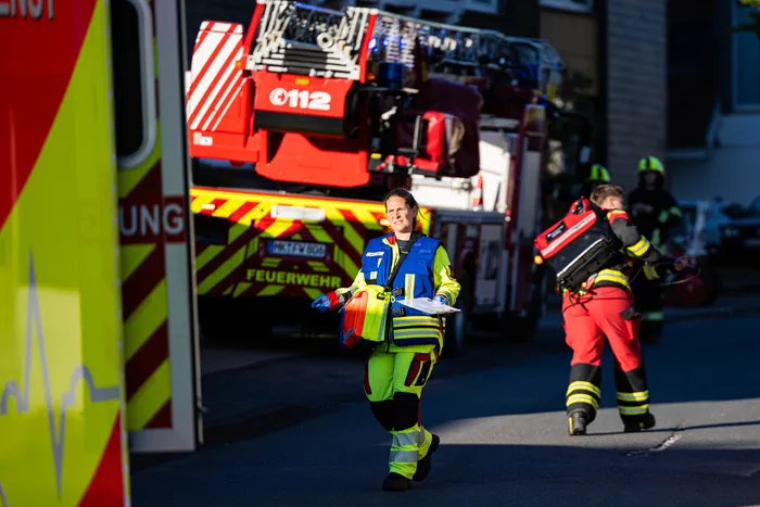 Eine Feuerwehrfrau in Einsatzkleidung geht mit einem Tablet vor einem roten Feuerwehrauto. Im Hintergrund sind weitere Einsatzkräfte und ein verrauchtes Gebäude zu sehen.