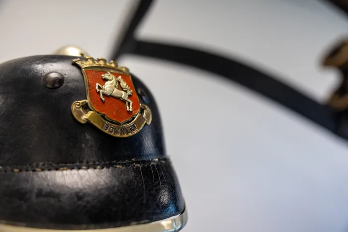 Ein schwarzer Feuerwehrhelm mit goldenen Verzierungen und einem Wappen, das einen springenden weißen Pferd auf rotem Grund zeigt. Darunter der Schriftzug 