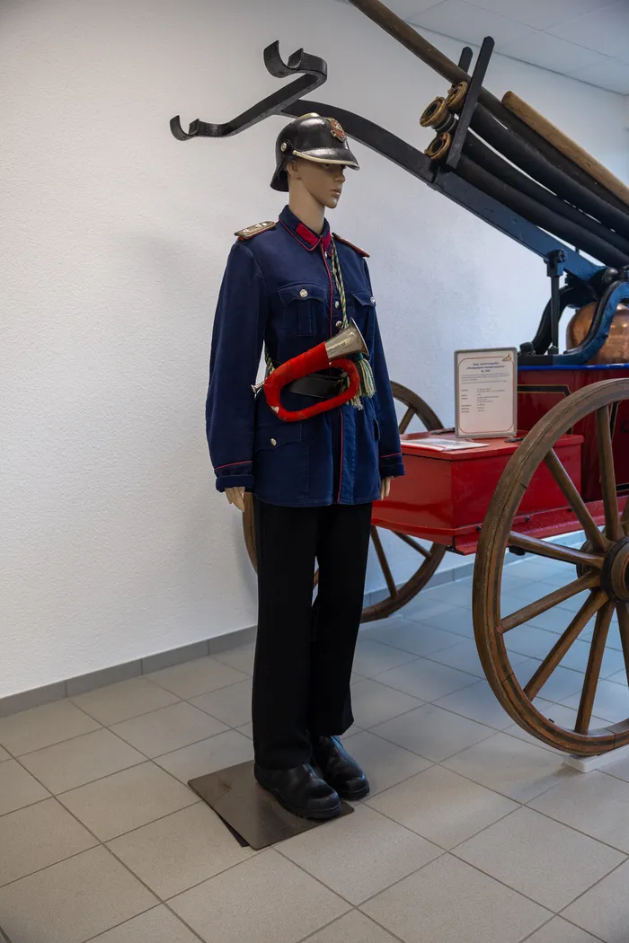 Eine Schaufensterpuppe trägt eine historische Feuerwehruniform mit Helm, Jacke, Hose und Stiefeln. Im Hintergrund steht ein roter, hölzerner Feuerwagen.