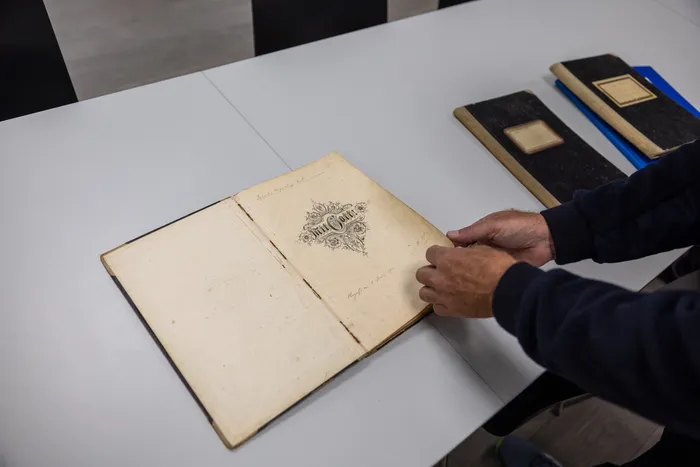 Ein Mann blättert in einem alten, geöffneten Buch mit vergilbten Seiten. Daneben liegen weitere historische Dokumente oder Fotoalben auf einem weißen Tisch.
