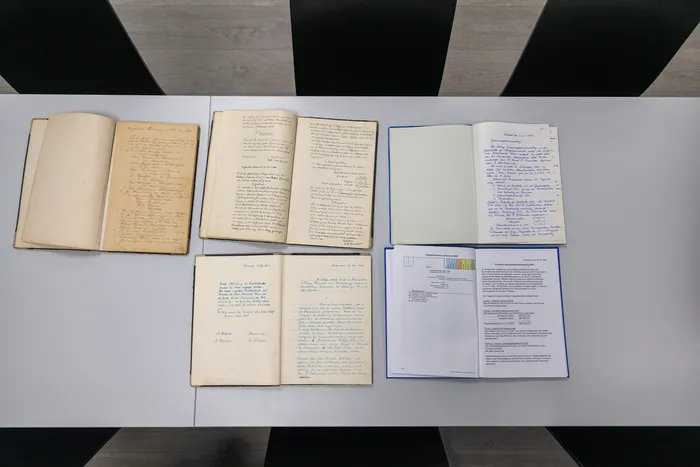 Mehrere geöffnete Notizbücher und Dokumente liegen auf einem hellgrauen Untergrund. Die Seiten sind mit handschriftlichen Notizen und Texten gefüllt.