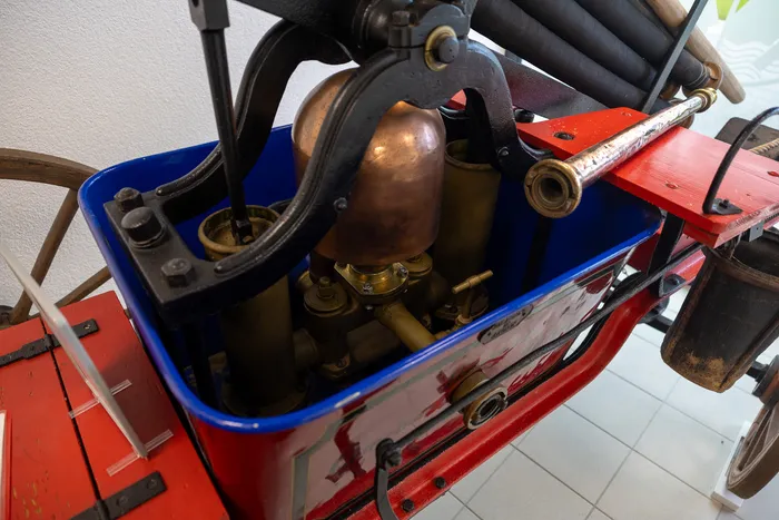 Nahaufnahme des Motors einer historischen Brandspritze, bestehend aus Kupferkessel, Rohren und Messingarmaturen. Das rot-blau lackierte Gehäuse zeigt eine stilisierte Darstellung einer Feuerspritze.