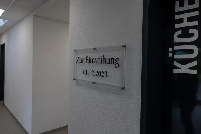 Ein transparentes Schild an einer hellen Wand kündigt die Einweihung am 01.12.2023 an. Rechts davon ist ein schmaler, schwarzer Streifen mit der Aufschrift „KÜCHE“ zu sehen.