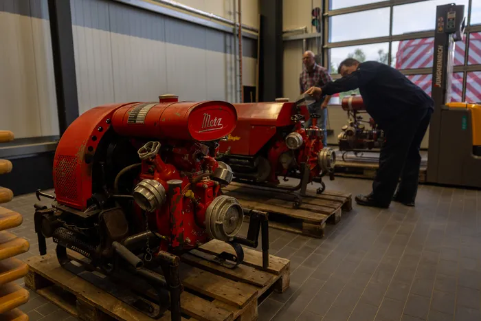 Ein rot lackierter Feuerlöschpumpenmotor steht auf Paletten im Vordergrund. Im Hintergrund arbeitet ein Mann an einem weiteren roten Gerät, während ein zweiter Mann zusieht.