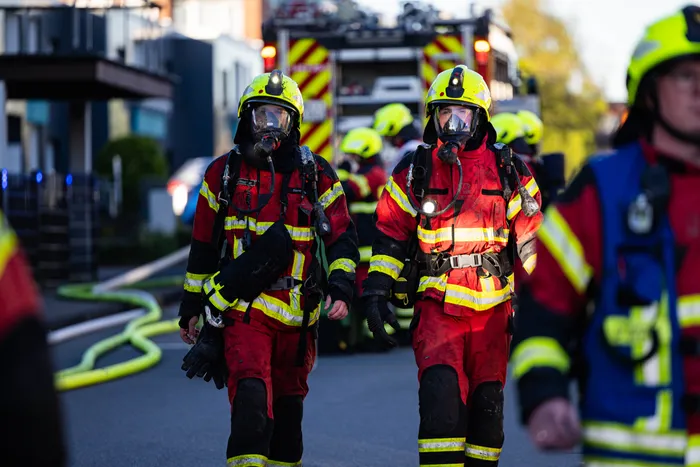 Zwei Feuerwehrleute mit Atemschutzmasken und Einsatzkleidung stehen vor einem Feuerwehrauto. Im Hintergrund sind weitere Einsatzkräfte zu sehen.