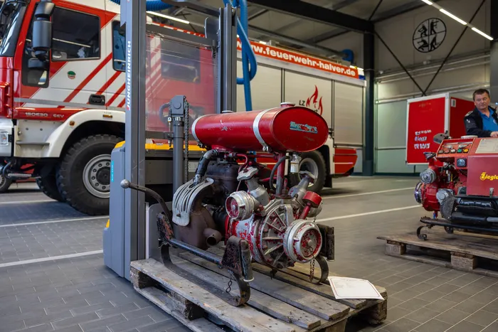 Roter Feuerwehrauto im Hintergrund, im Vordergrund ein zerlegter Motor auf einer Palette. Im Hintergrund weitere Fahrzeuge und ein Mann in Uniform.
