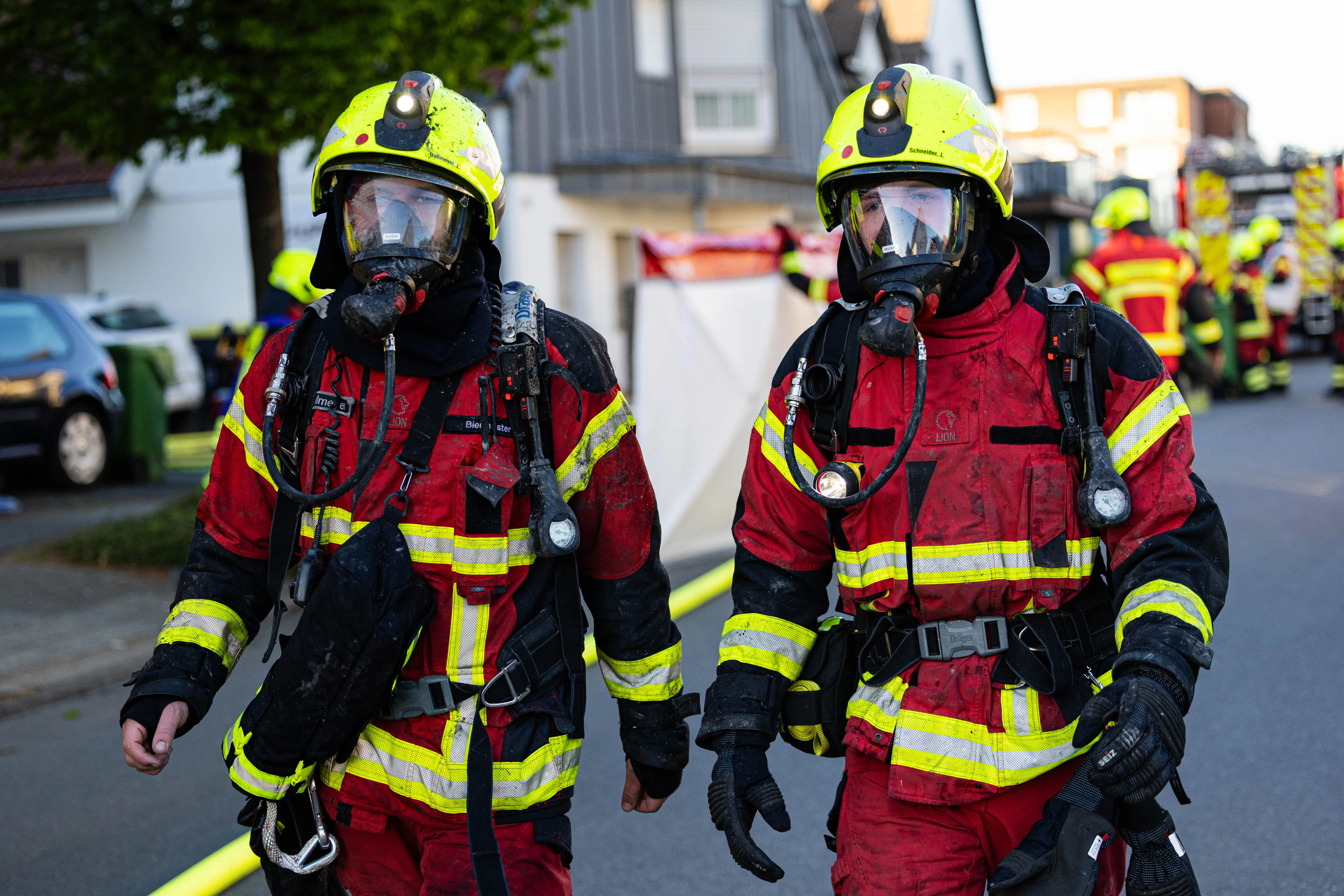 Zwei Feuerwehrleute mit Atemschutzmasken und Einsatzkleidung stehen vor einer Straße mit Einsatzfahrzeugen. Im Hintergrund ist ein Wohnhaus zu sehen.