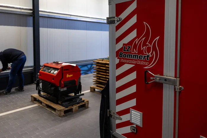 Rote Feuerwehrausrüstung steht auf einer Palette in einer Lagerhalle, während im Hintergrund eine Person zu sehen ist. Rechts ist die rote Tür eines Feuerwehrautos mit dem Schriftzug 'LZ Bommert Feuerwehr Halver' zu erkennen.