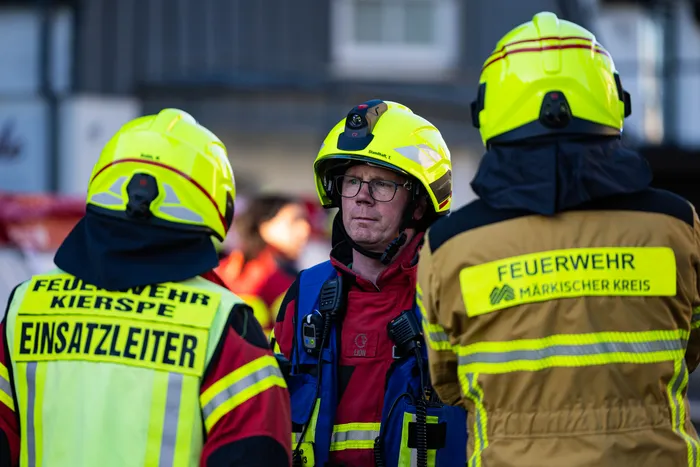 Ein Feuerwehrmann mit Brille und rotem Schutzanzug blickt nachdenklich in die Ferne, flankiert von zwei weiteren Feuerwehrleuten mit gelben Helmen. Auf den Westen der Kollegen sind die Aufschriften „FEUERWEHR KIERSPE EINSATZLEITER“ und „FEUERWEHR MÄRKISCHER KREIS“ zu sehen.