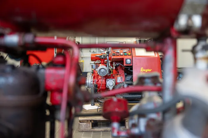 Roter Feuerwehrmotor mit dem Schriftzug „Ziegler“ im Fokus, umgeben von roten Stahlträgern. Das Bild zeigt Details der Technik eines Feuerwehrautos.