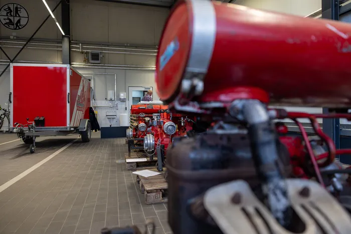 Roter Oldtimer-Feuerwehrauto steht in einer Werkstatt auf Paletten, während ein Mann im Hintergrund arbeitet. Im Hintergrund ein roter Anhänger und ein Logo an der Decke.