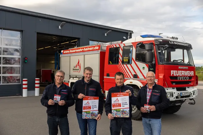 Vier Feuerwehrleute in Uniformen stehen vor einem Feuerwehrauto und halten Einladungen zum Tag der offenen Tür hoch. Im Hintergrund ist die Fahrzeughalle der Feuerwache zu sehen.
