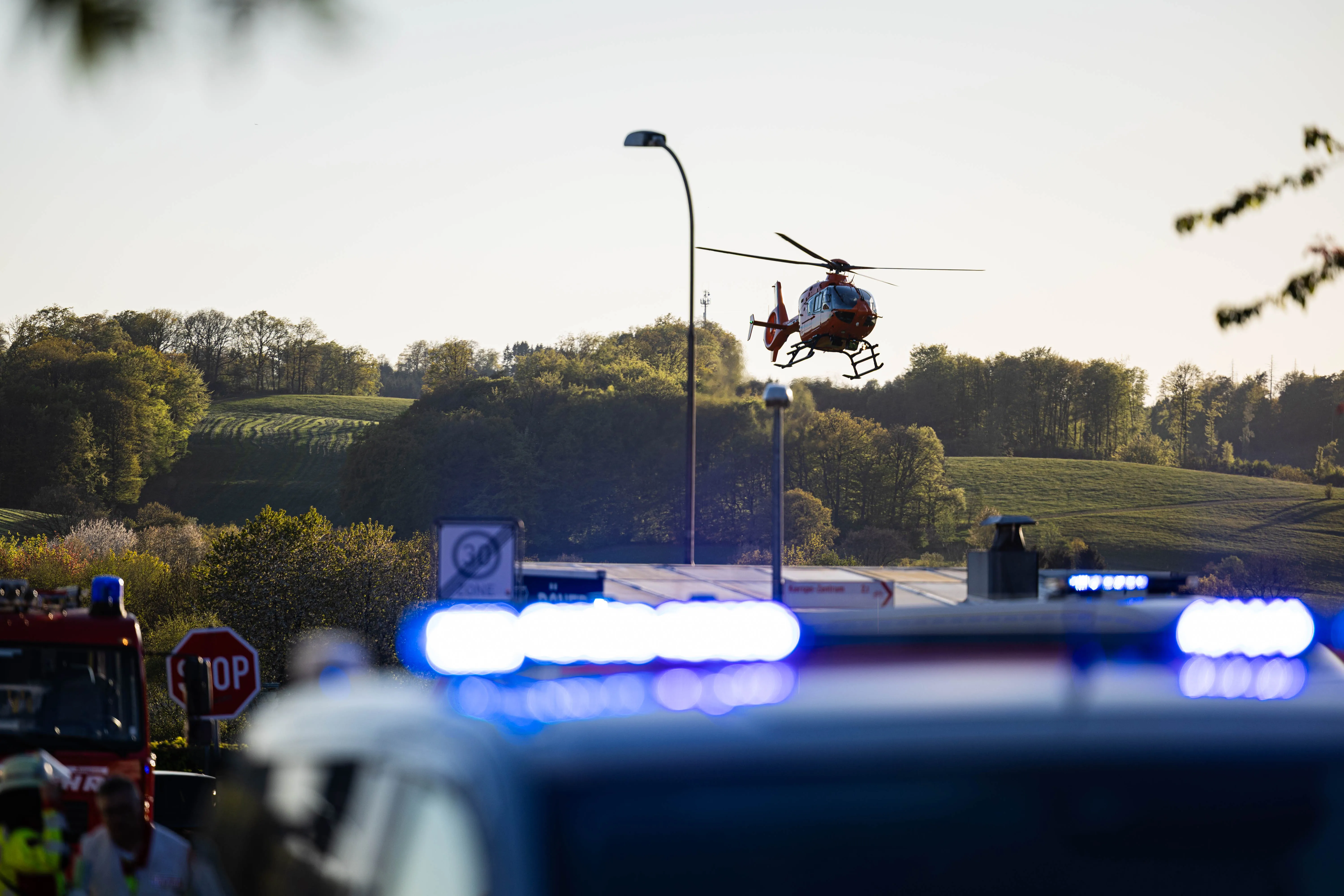 Ein Rettungshubschrauber landet auf einer Straße, während im Hintergrund Einsatzfahrzeuge mit Blaulicht zu sehen sind. Der Einsatzort ist ein Wohnungsbrand in Kierspe, bei dem eine junge Frau ums Leben kam.
