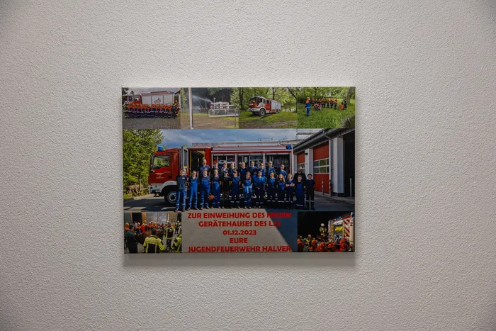 Ein gerahmtes Foto zeigt die Jugendfeuerwehr Halver vor einem roten Feuerwehrauto und weiteren Bildern von Feuerwehrleuten und Fahrzeugen. Der Text auf dem Foto erinnert an die Einweihung des Gerätehauses am 01.12.2023.