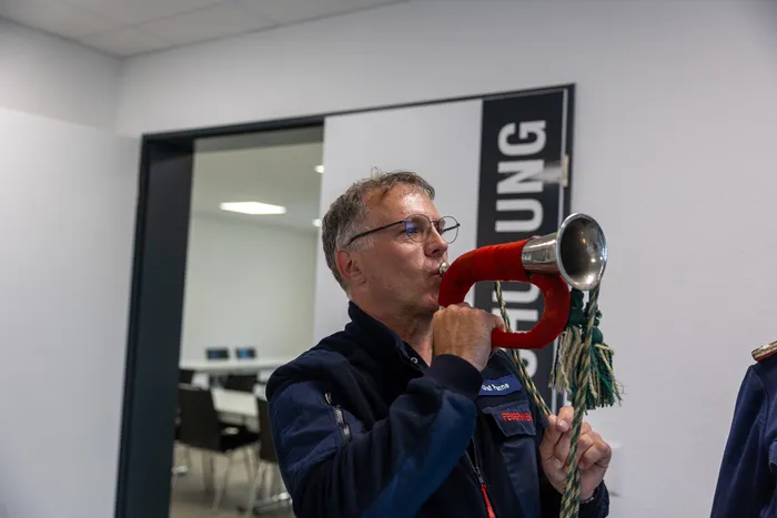 Ein Mann mit Brille bläst in ein goldenes Horn. Im Hintergrund ist ein großes Banner mit der Aufschrift „FEUERWEHR“ zu sehen.