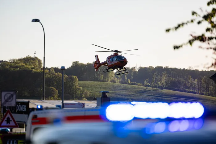Rettungshubschrauber landet neben Einsatzfahrzeugen mit Blaulicht vor dem Hintergrund von Bäumen und Hügeln, nachdem bei einem Wohnungsbrand in Kierspe vier Personen schwer verletzt wurden.
