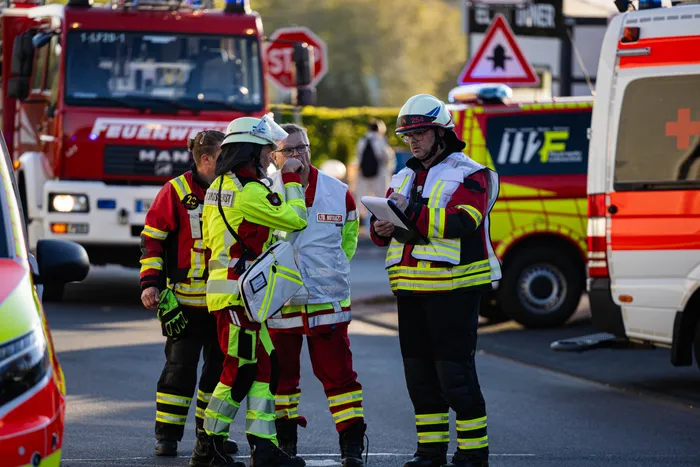 Mehrere Einsatzkräfte mit Schutzhelmen und Warnwesten besprechen Notizen an einem Einsatzort mit mehreren Feuerwehrfahrzeugen und einem Rettungswagen im Hintergrund.