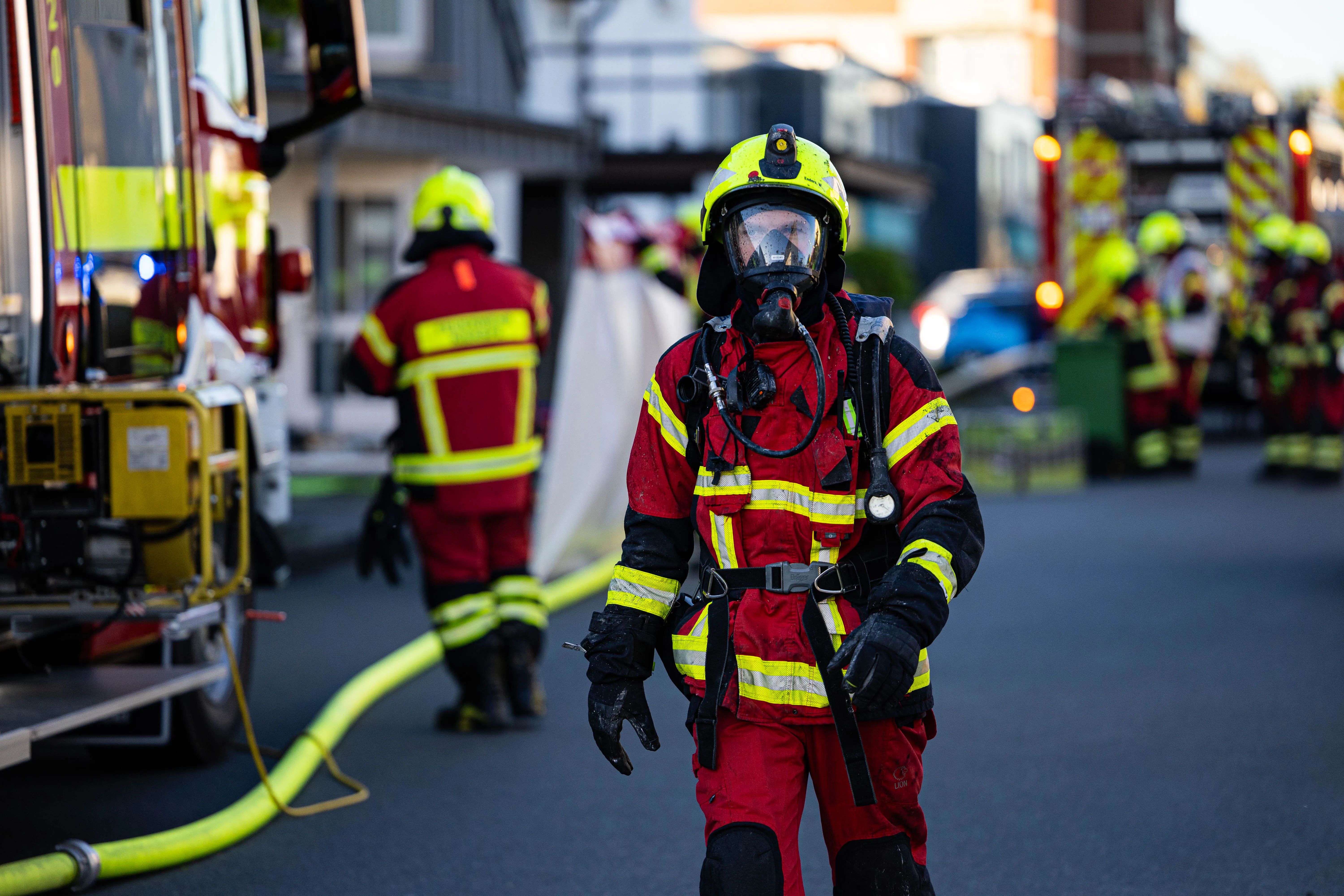 Ein Feuerwehrmann in voller Schutzausrüstung steht im Vordergrund, während im Hintergrund weitere Einsatzkräfte und Fahrzeuge zu sehen sind. Der Einsatz erfolgte nach einem tödlichen Wohnungsbrand in Kierspe.