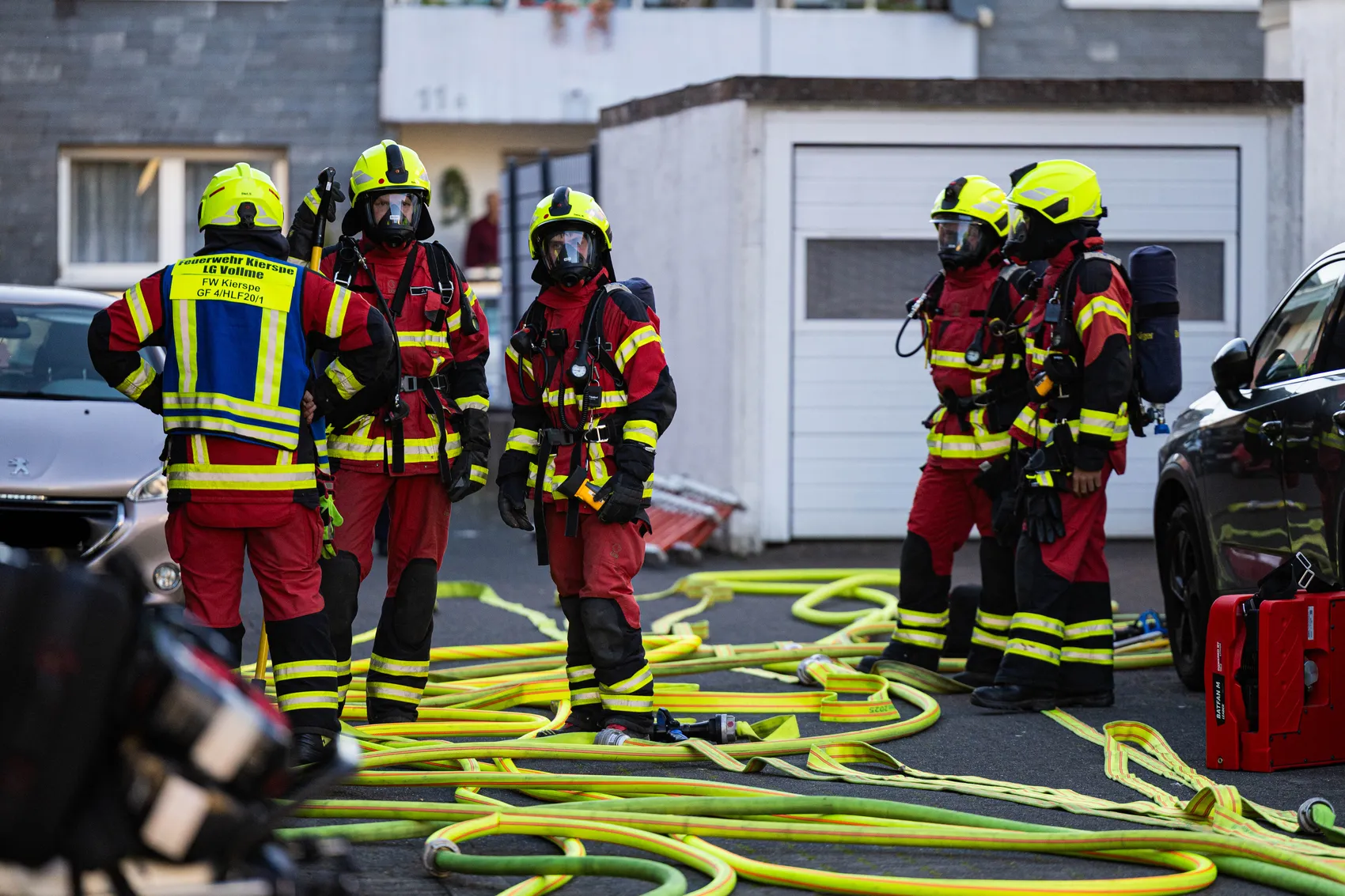 Feuerwehrleute in voller Montur stehen vor einem Mehrfamilienhaus mit Brandspuren, während ein gelber Schlauch vor ihnen liegt. Im Hintergrund sind geparkte Autos zu sehen.