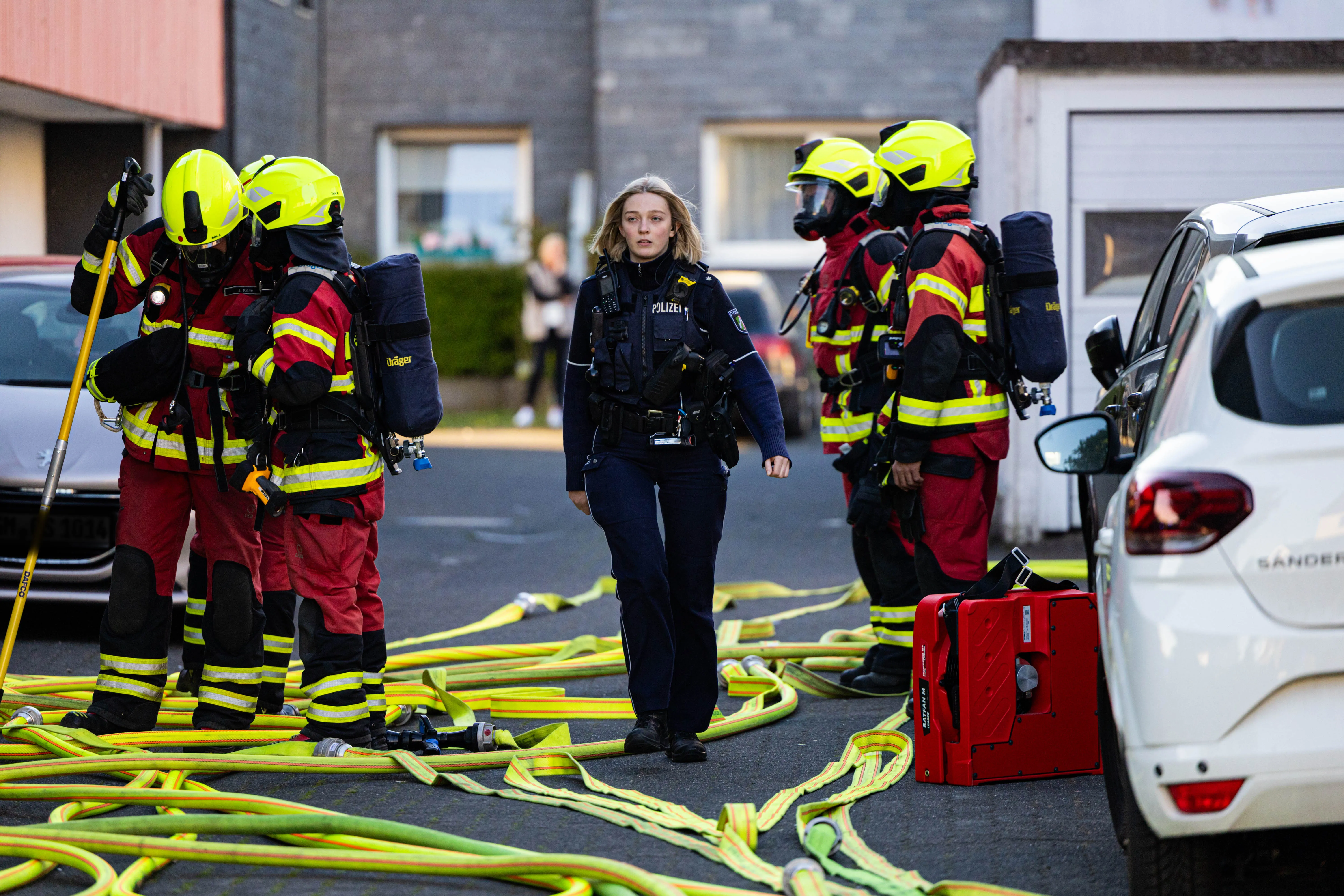 Feuerwehrleute in voller Montur und eine Polizistin stehen vor einem Gebäude, umgeben von Löschschläuchen. Im Hintergrund ist ein weißer PKW zu sehen.