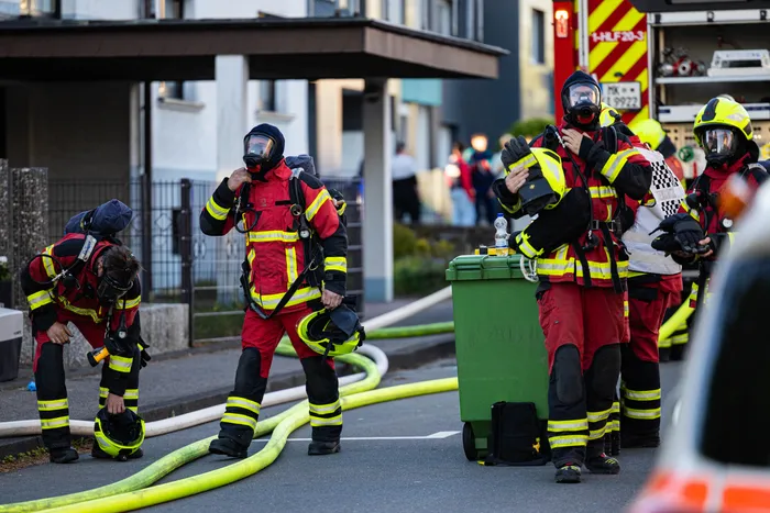 Feuerwehrleute in voller Montur stehen und arbeiten an einem Einsatzort vor einem Wohnhaus nach einem Brand. Im Vordergrund liegen Schläuche und ein grüner Müllcontainer.