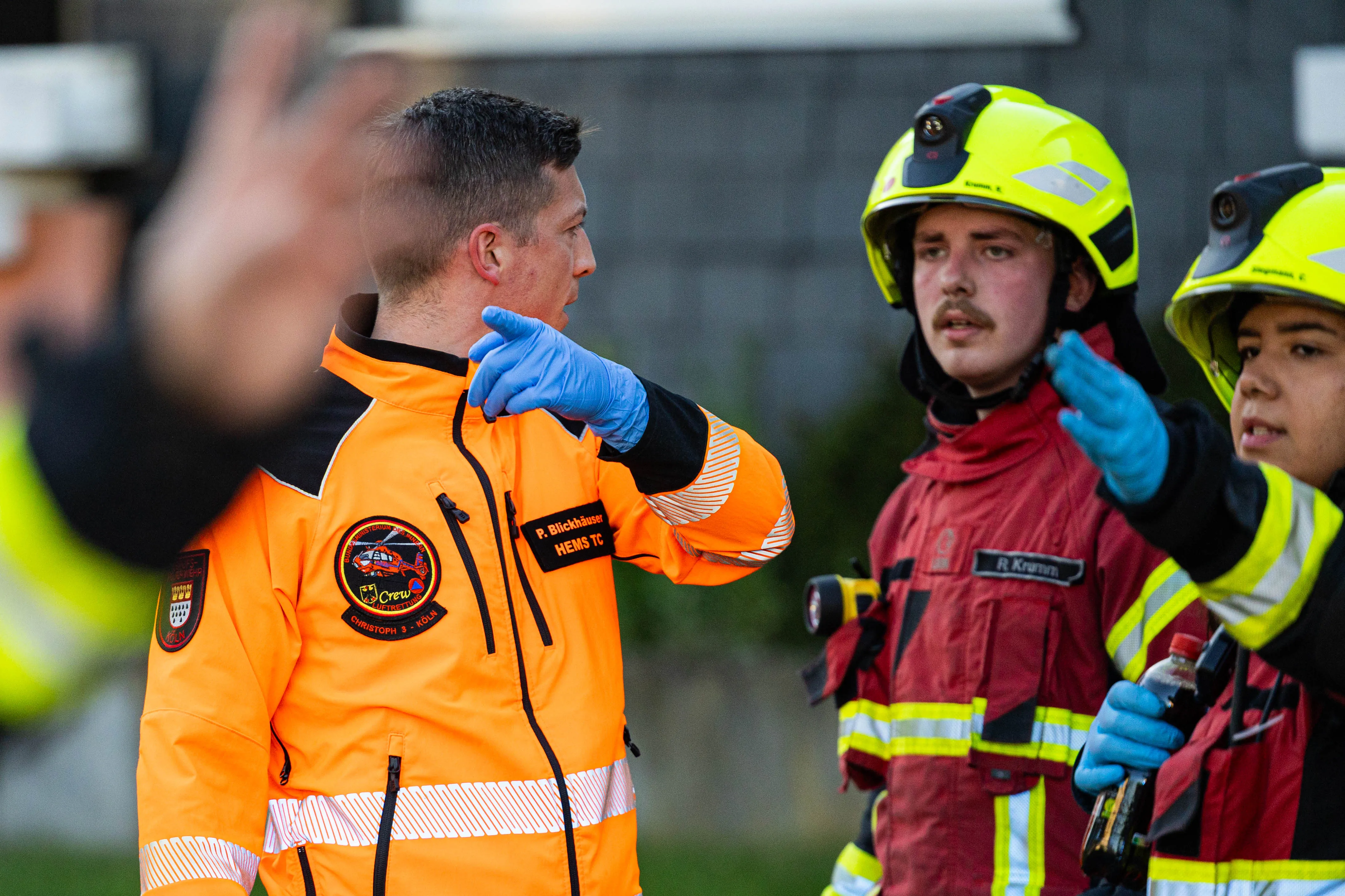 Ein Rettungssanitäter in orangefarbener Jacke mit Handschuhen spricht in Kierspe mit zwei Feuerwehrleuten in voller Montur, während im Hintergrund weitere Einsatzkräfte zu sehen sind.