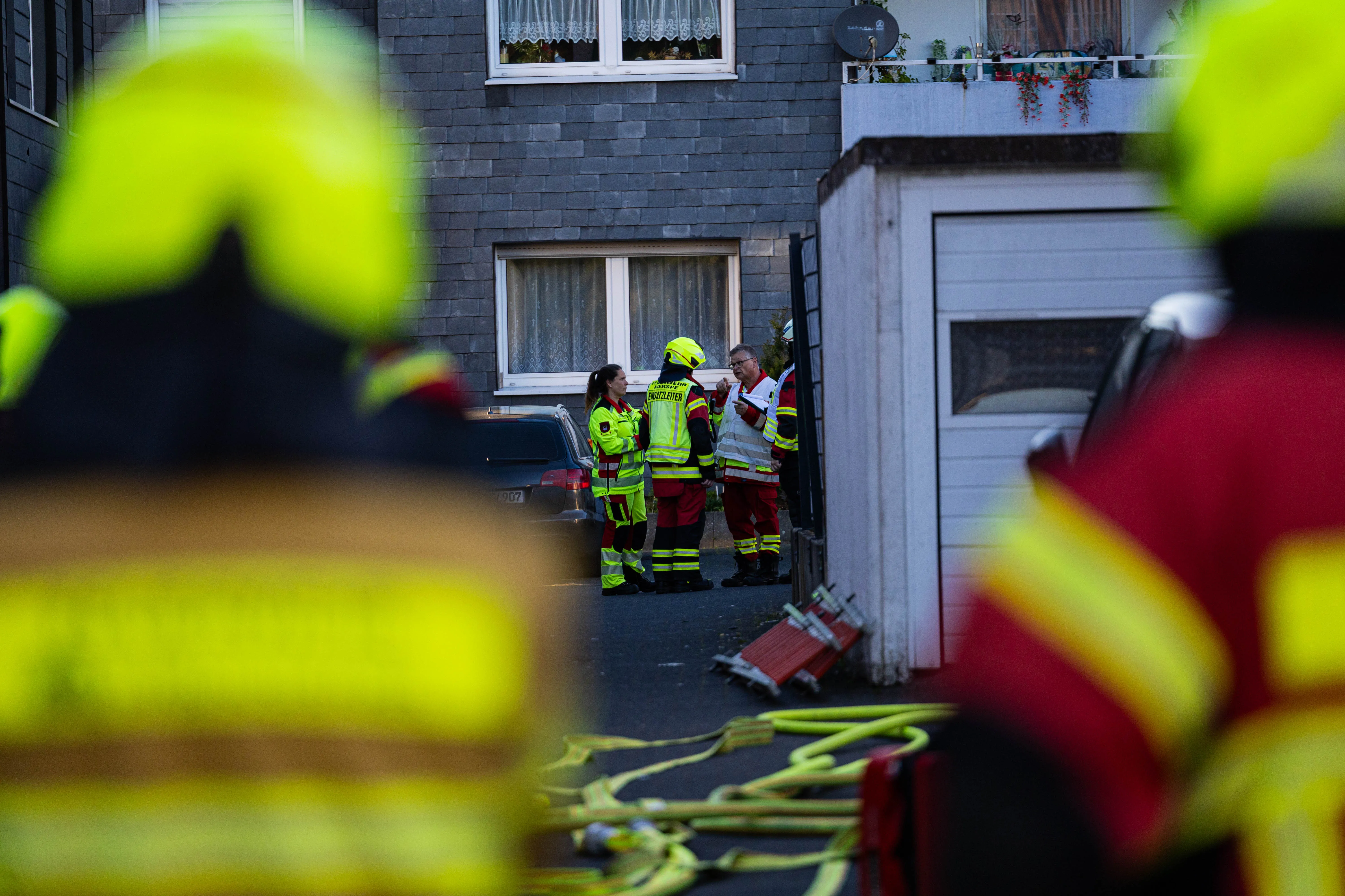 Feuerwehrleute stehen vor einem Wohnhaus, während Schläuche und Geräte bereitliegen. Im Hintergrund ist ein Auto geparkt und ein Balkon mit Blumenkästen zu sehen.