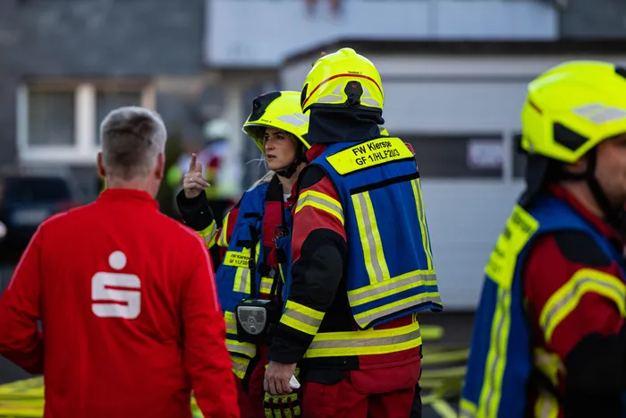 Feuerwehrleute in Kierspe besprechen während eines Wohnungsbrandes Einsatzmaßnahmen. Im Hintergrund sind weitere Einsatzkräfte und ein verrauchter Gebäudeteil zu sehen.