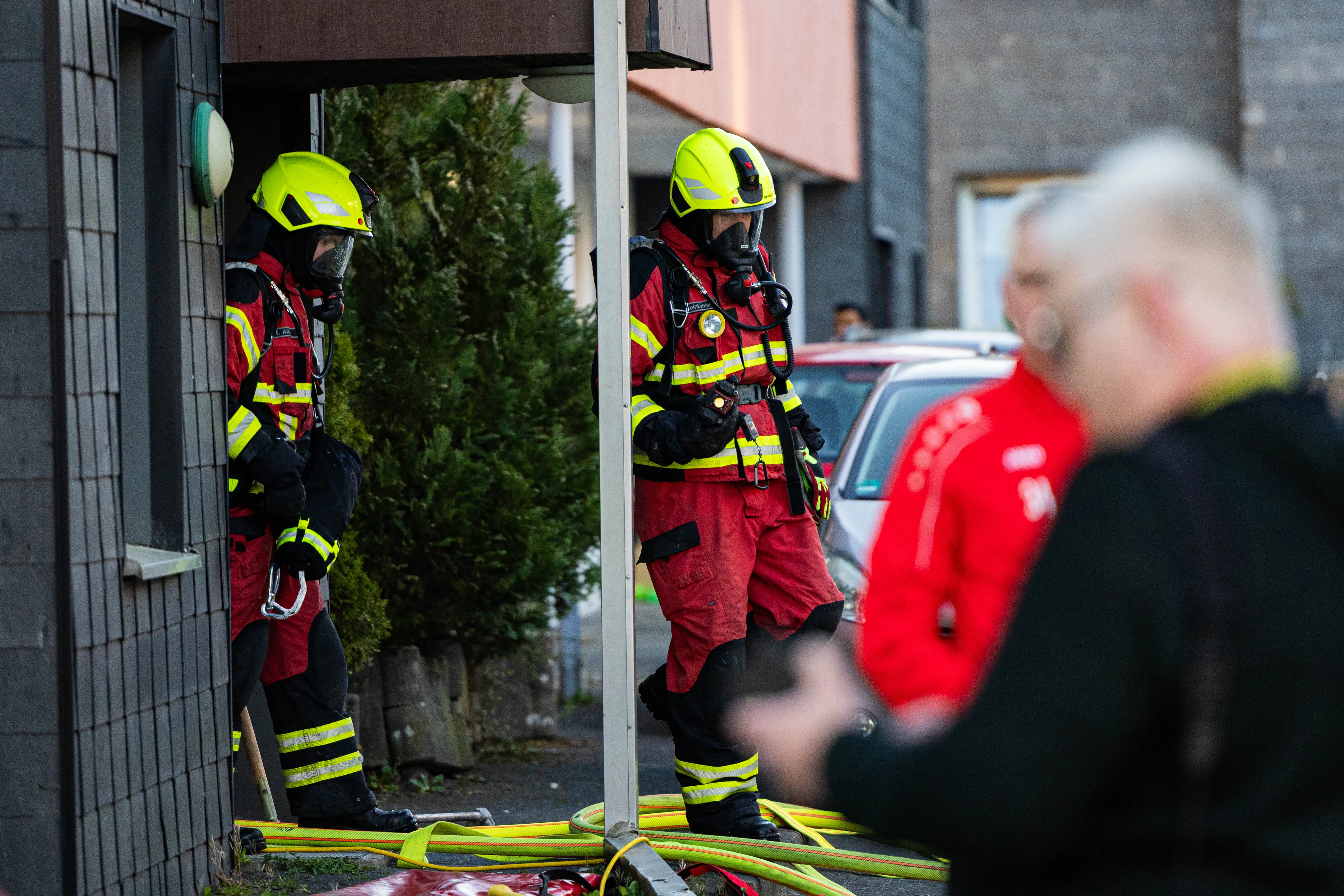 Zwei Feuerwehrleute in voller Montur stehen vor einem Gebäude, während im Vordergrund ein Schlauch und eine verschwommene Person zu sehen sind. Im Hintergrund sind weitere Einsatzkräfte und Fahrzeuge erkennbar.