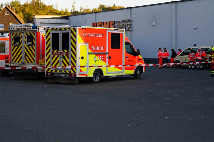 Zwei Rettungswagen mit Aufschrift „Rettungsdienst“ und „Notruf 112“ stehen auf einem Parkplatz vor einem Gebäude. Im Hintergrund sind Einsatzkräfte und eine Absperrung zu sehen.