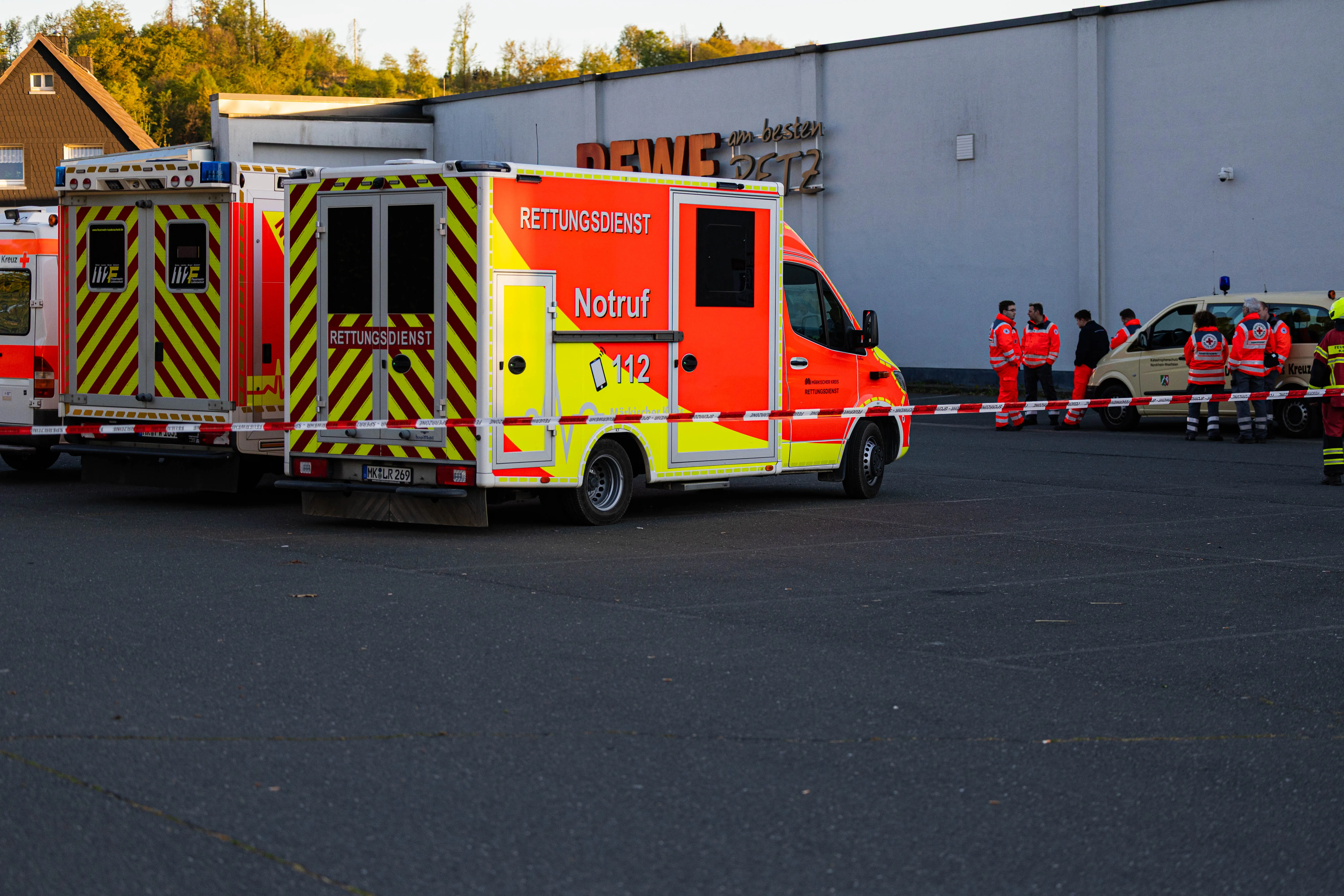 Zwei Rettungswagen mit Aufschrift „Rettungsdienst“ und „Notruf 112“ stehen vor einem Gebäude, während im Hintergrund Einsatzkräfte an einer Absperrung stehen.