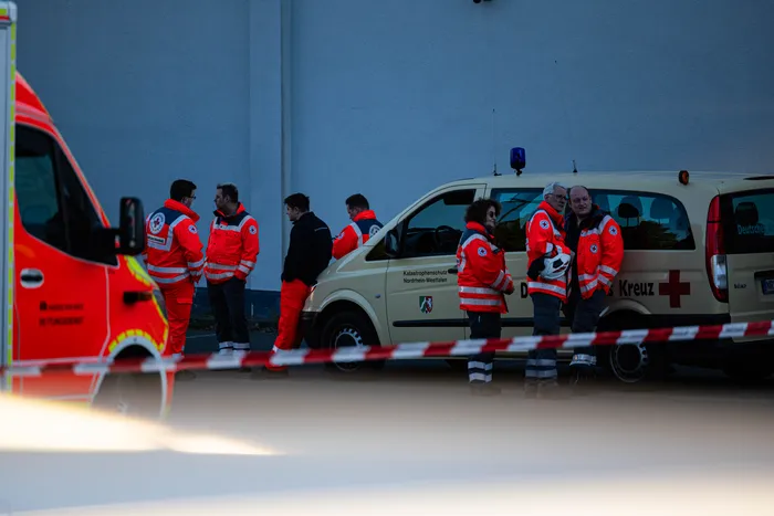 Rettungskräfte des Deutschen Roten Kreuzes und des THW vor einem beigefarbenen Mannschaftswagen an der Einsatzstelle eines Wohnungsbrandes, abgesperrt durch rotes Absperrband.