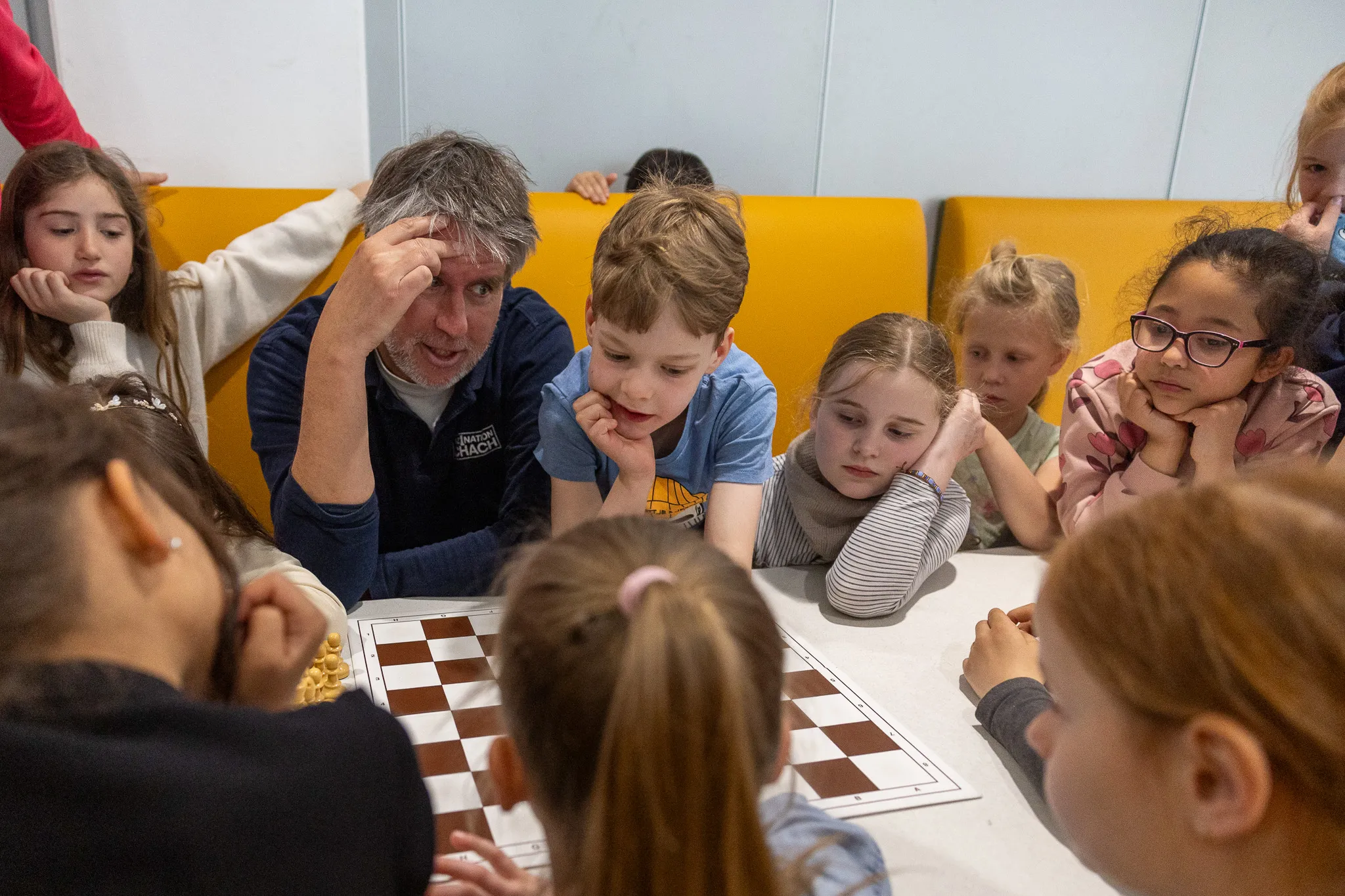 Schachgroßmeister Sebastian Siebrecht erklärt Kindern an einem Schachbrett den nächsten Zug. Die Schülerinnen und Schüler sitzen konzentriert um den Tisch.