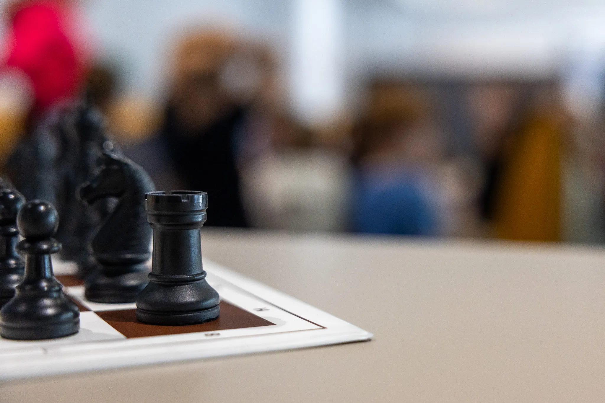 Schwarze Schachfiguren auf einem Schachbrett im Vordergrund, im Hintergrund verschwommene Aufnahme von Menschen in einem Raum.