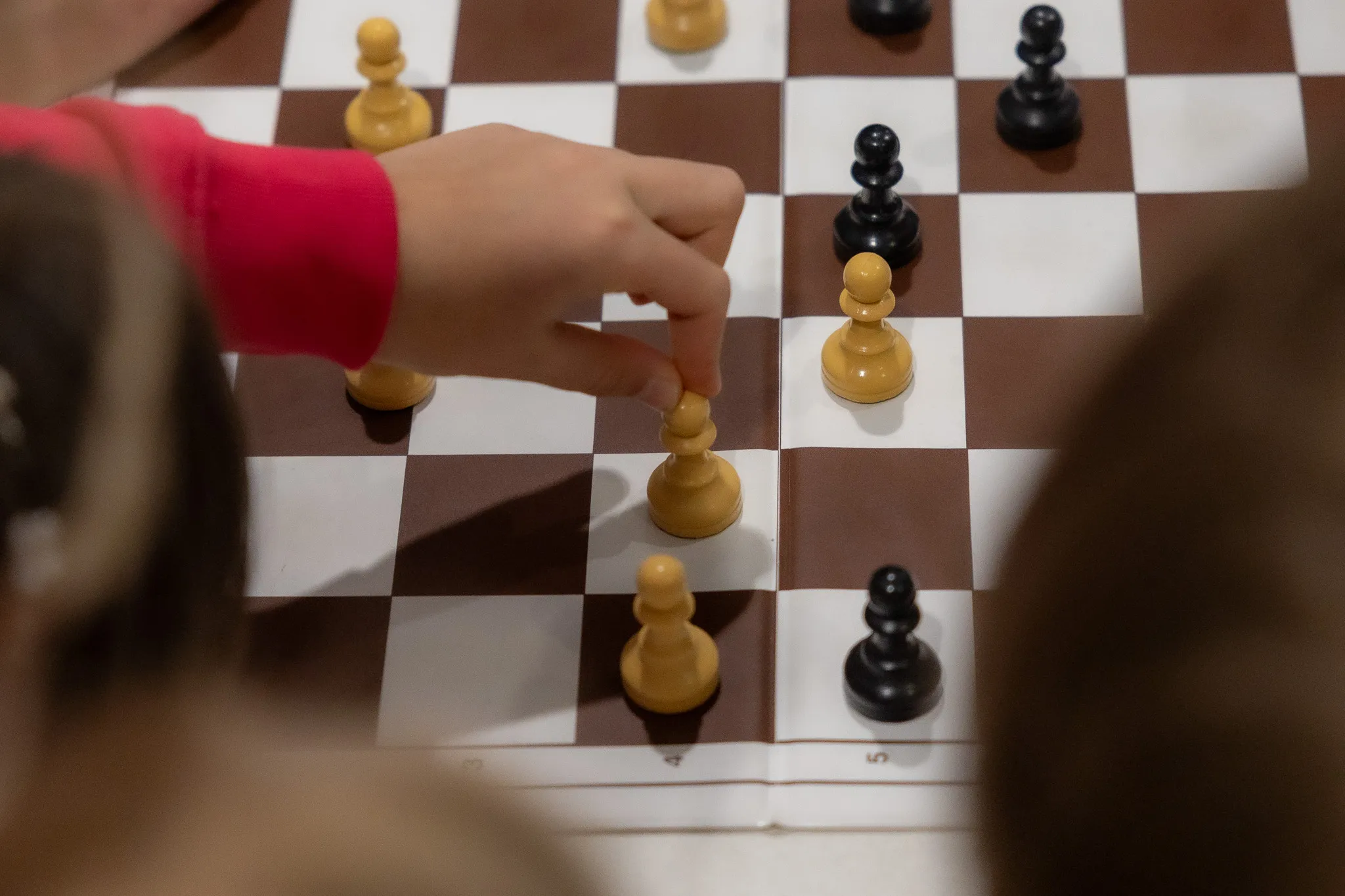 Kind verschiebt eine weiße Schachfigur auf dem Schachbrett, während ein weiterer Spieler im Hintergrund sitzt.