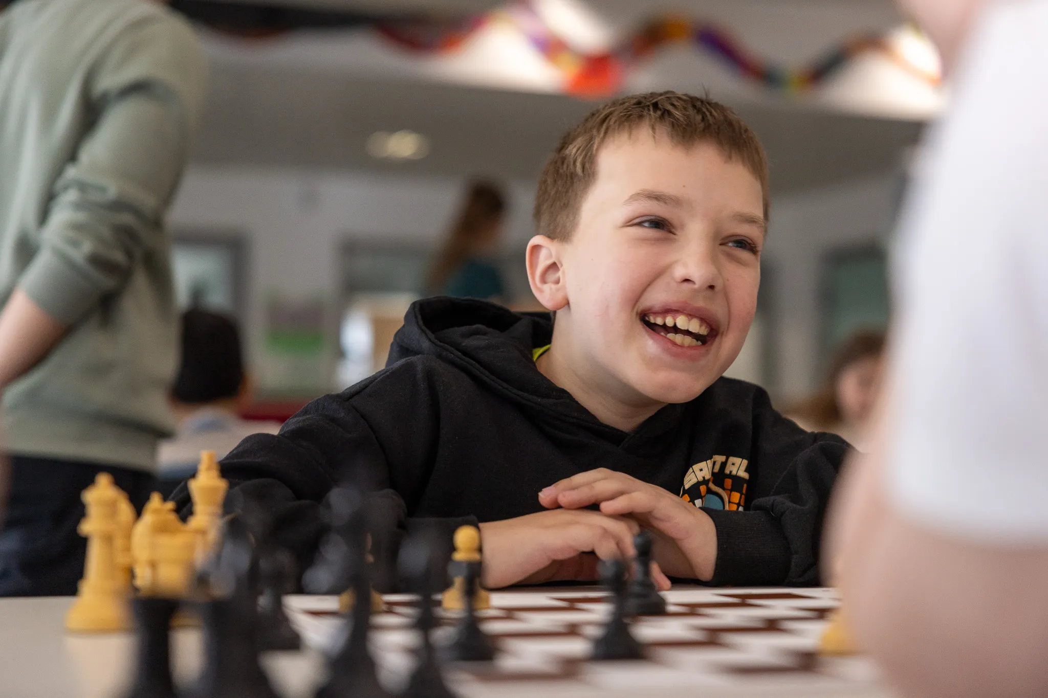 Junge lacht beim Schachspielen mit schwarzen und weißen Figuren. Im Hintergrund sind weitere Kinder und ein Schachgroßmeister zu sehen.