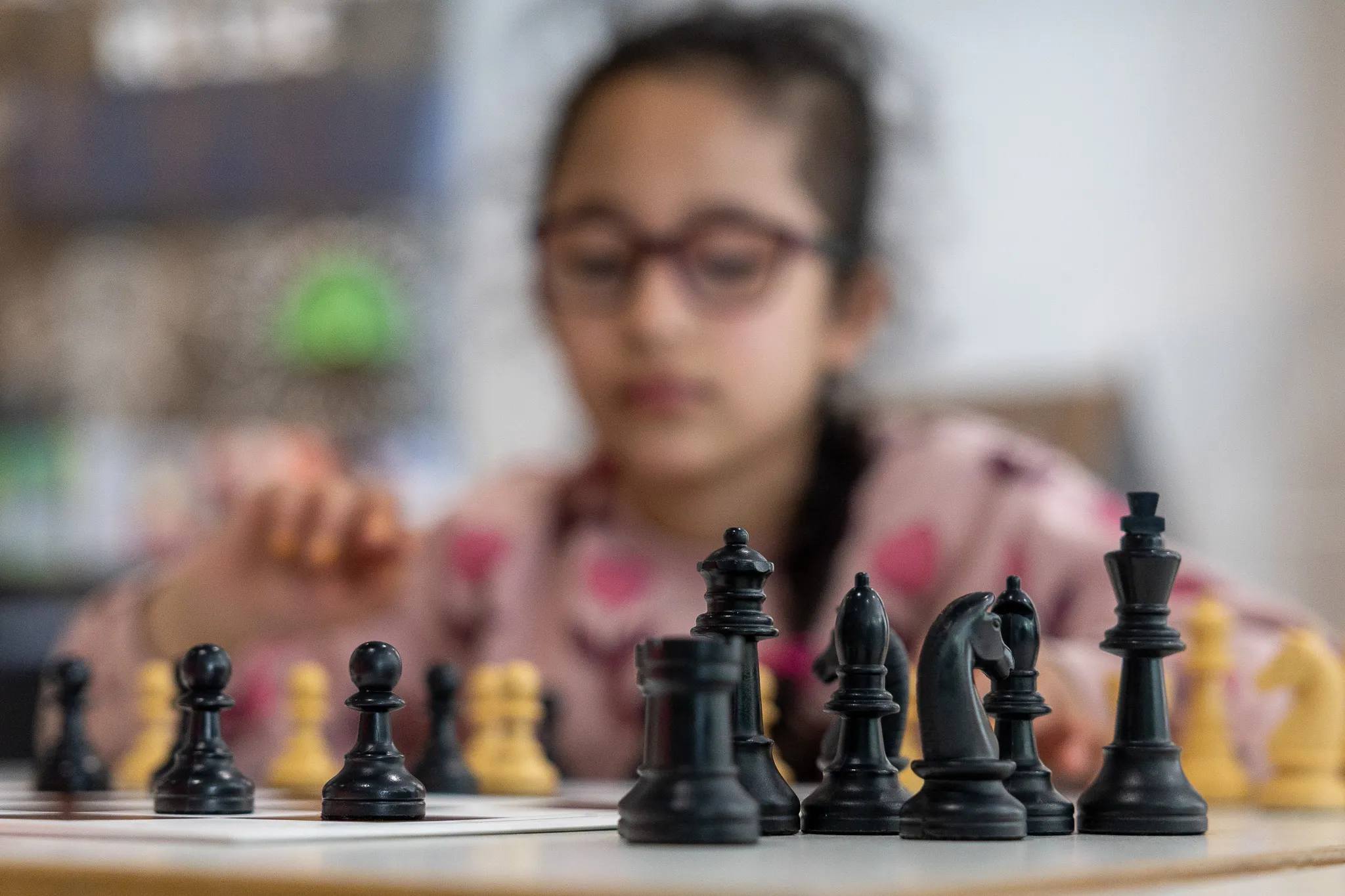 Schachfiguren stehen im Vordergrund, im Hintergrund ein Mädchen mit Brille, das konzentriert auf das Spiel blickt.