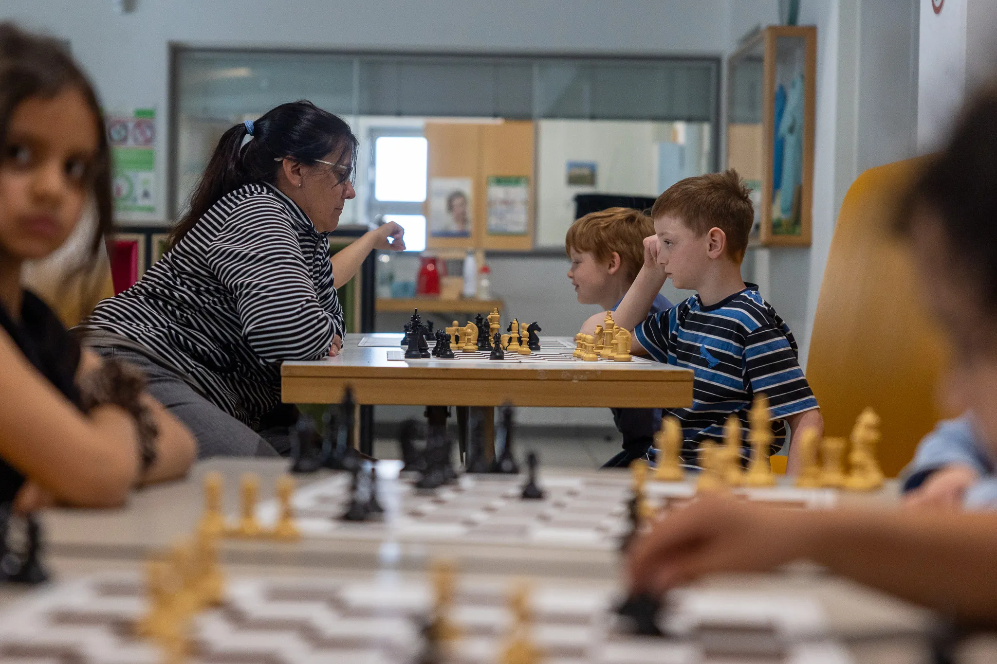 Zwei Kinder spielen Schach unter Anleitung einer Frau, während andere Kinder im Hintergrund zusehen. Im Hintergrund sind Schränke und eine Tür zu sehen.
