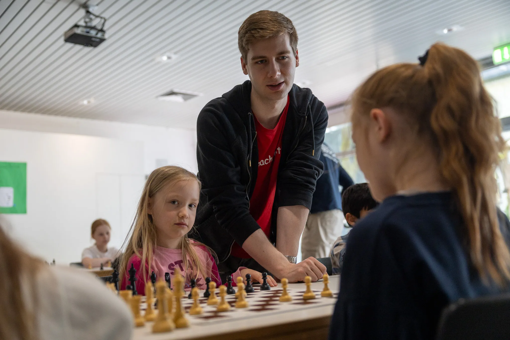 Ein junger Mann erklärt einem Mädchen und einem Jungen die Züge auf einem Schachbrett. Im Hintergrund sitzen weitere Kinder an Tischen.
