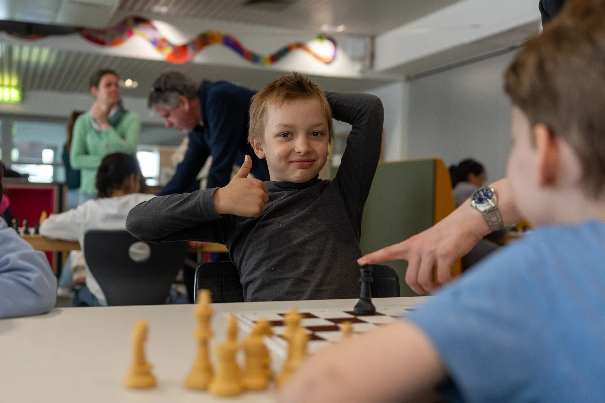 Junge mit rotem Haar zeigt Daumen hoch, während er Schach spielt. Im Hintergrund weitere Kinder und ein Mann, vermutlich Schachgroßmeister Sebastian Siebrecht.