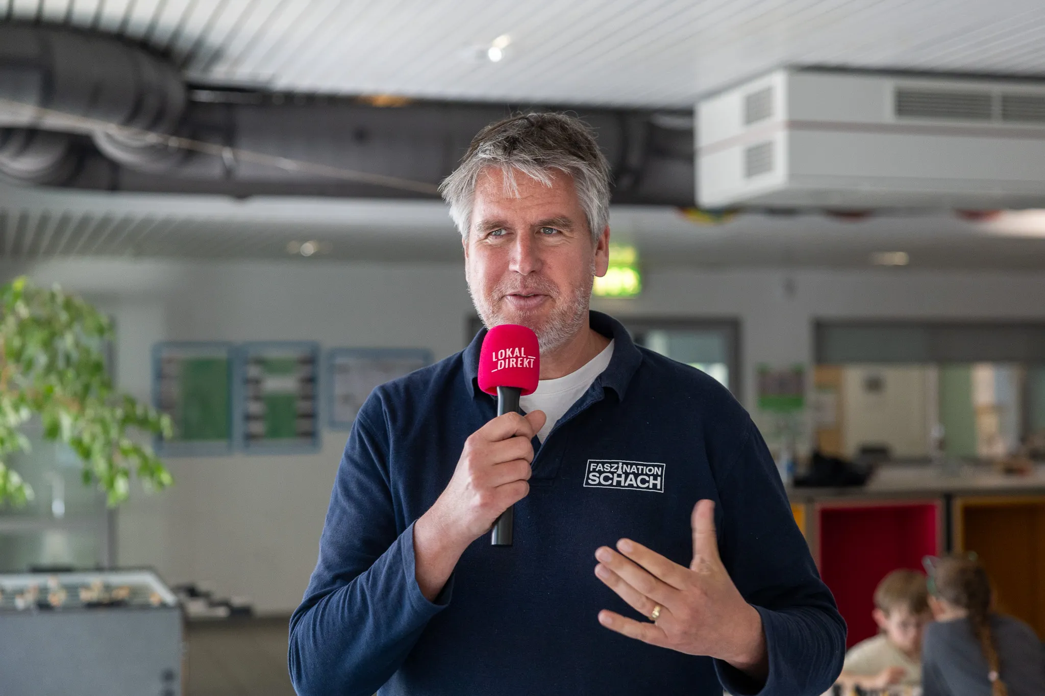 Schachgroßmeister Sebastian Siebrecht hält ein Mikrofon mit dem Logo „LokalDirekt“ und spricht in einem Innenraum.